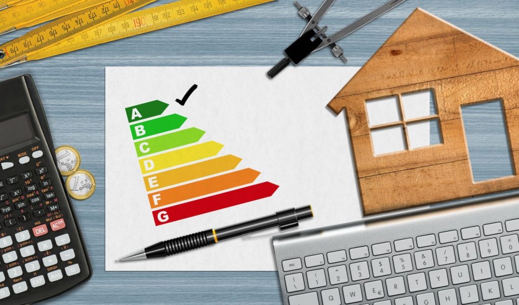 Ocena efektywności energetycznej domu. Mały drewniany model domu na biurku z wykresem efektywności energetycznej, kalkulatorem, składaną linijką, kompasem, ołówkiem i klawiaturą komputera oraz miejscem do kopiowania.