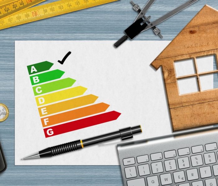 Ocena efektywności energetycznej domu. Mały drewniany model domu na biurku z wykresem efektywności energetycznej, kalkulatorem, składaną linijką, kompasem, ołówkiem i klawiaturą komputera oraz miejscem do kopiowania.