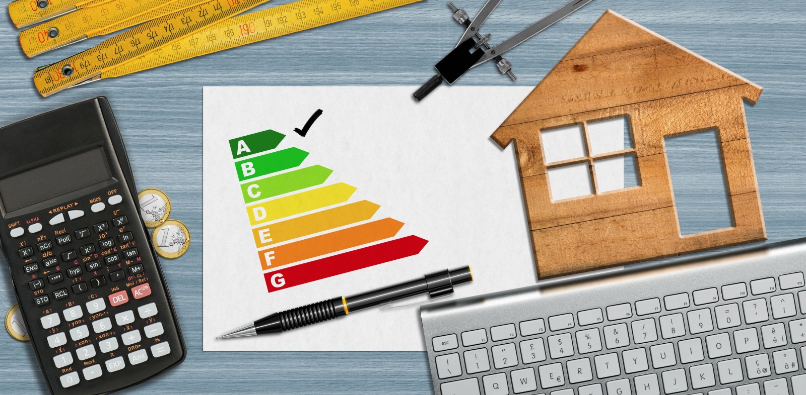 Ocena efektywności energetycznej domu. Mały drewniany model domu na biurku z wykresem efektywności energetycznej, kalkulatorem, składaną linijką, kompasem, ołówkiem i klawiaturą komputera oraz miejscem do kopiowania.