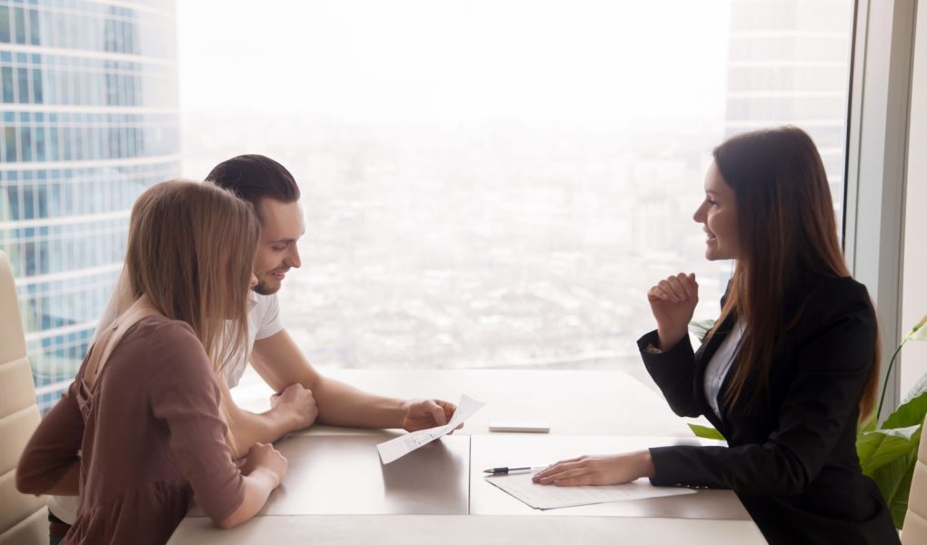 Widok z boku na pośrednika i młodą parę siedzącą przy biurku i omawiającą nieruchomość na sprzedaż. Potencjalni nabywcy trzymający plan domu, rozważający inwestycję w dom lub zaciągnięcie kredytu na zakup nieruchomości