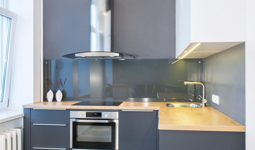 Nowoczesne meble kuchenne we wnętrzu mieszkania z drewnianymi akcentami. Oświetlenie ledowe. Europejski, skandynawski styl. Malowane szare fasady. Szklany panel ścienny. Zarządzanie projektem. Nieruchomości. Wynajem. Sprzedaż.