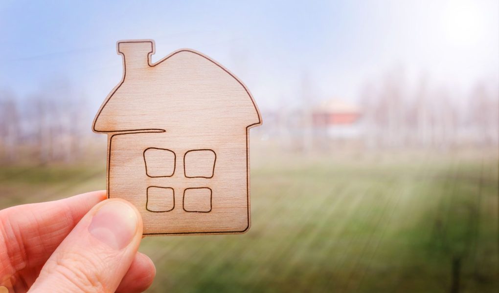 Model domu w ręku agenta nieruchomości lub osoby, która marzy o zakupie swojego domu na tle nieba i łąki z kopią przestrzeni. Oferta zakupu domu, ubezpieczenie nieruchomości, koncepcja przystępnych cenowo mieszkań.