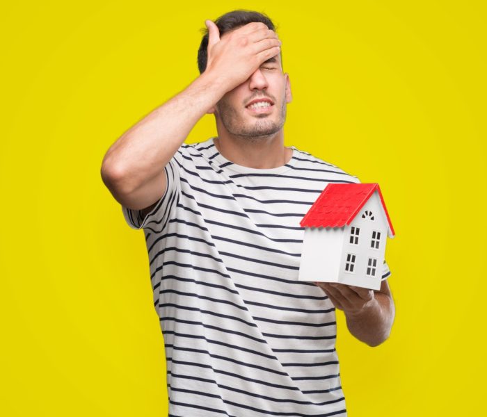 Przystojny agent nieruchomości trzymający dom zestresowany z ręką na głowie, zszokowany ze wstydem i zaskoczeniem na twarzy, zły i sfrustrowany. Strach i zdenerwowanie z powodu błędu.