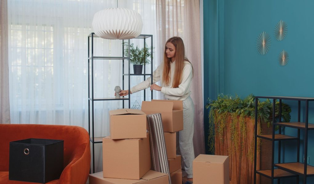 Kobieta wchodzi do pokoju trzymając kartonowe pudła, rozpakowując paczki z meblami po wynajęciu lub zakupie nowego mieszkania w domu. Dziewczyna skoncentrowana na przeprowadzce. Ludzie styl życia