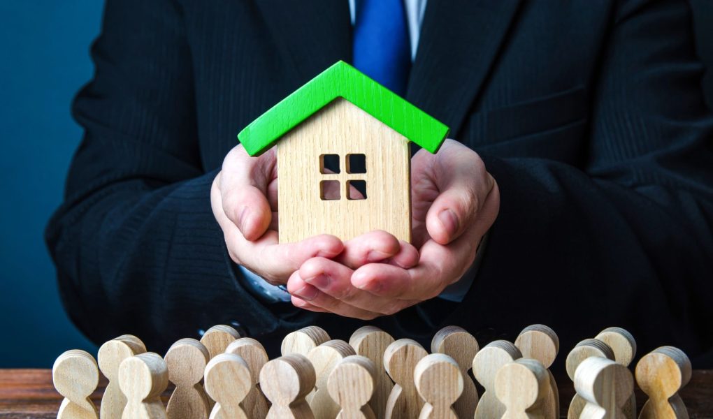 Mężczyzna prezentuje ludziom dom. Oferta przystępnego kredytu hipotecznego na zakup domu. Mieszkania gwarantowane przez państwo. Zapewnienie nieruchomości socjalnych dla osób potrzebujących. Wynajem mieszkań i domów.