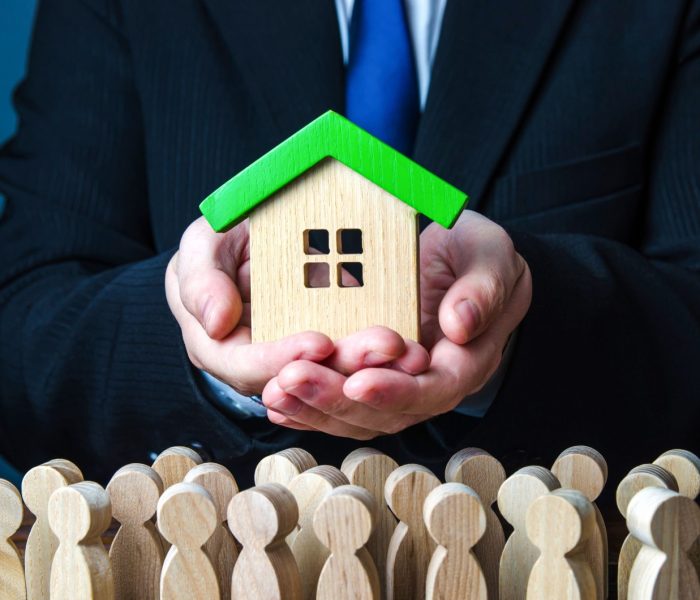 Mężczyzna prezentuje ludziom dom. Oferta przystępnego kredytu hipotecznego na zakup domu. Mieszkania gwarantowane przez państwo. Zapewnienie nieruchomości socjalnych dla osób potrzebujących. Wynajem mieszkań i domów.