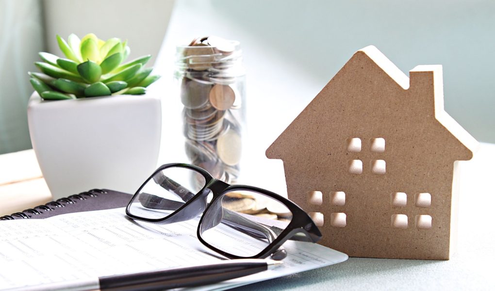 Biznes, finanse, oszczędzanie pieniędzy, bankowość, pożyczka na nieruchomość lub koncepcja kredytu hipotecznego: Drewniany model domu, monety, okulary i książeczka oszczędnościowa lub zestawienie finansowe na biurku