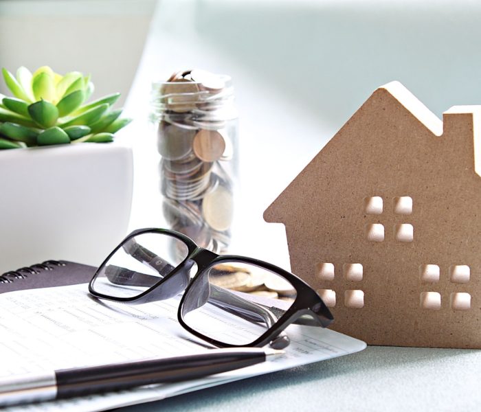 Biznes, finanse, oszczędzanie pieniędzy, bankowość, pożyczka na nieruchomość lub koncepcja kredytu hipotecznego: Drewniany model domu, monety, okulary i książeczka oszczędnościowa lub zestawienie finansowe na biurku