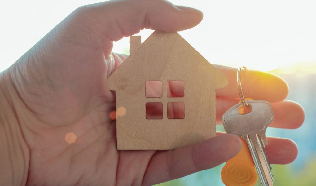 W ręku mężczyzny mały drewniany domek i klucz, na tle słońca w letni dzień. Koncepcja - kupowanie nieruchomości, marzenie o własnym domu, niedrogie mieszkania, sprzedaż nieruchomości.
