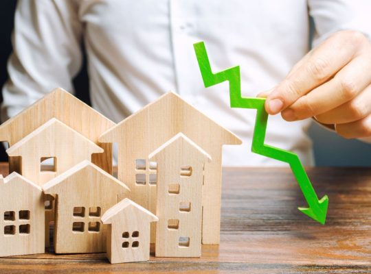 Analityk trzyma strzałkę w dół w pobliżu drewnianych domów. Koncepcja spadającego rynku nieruchomości. Niskie ceny i koszty mieszkań. Obniżone oprocentowanie kredytów hipotecznych i popyt na mieszkania. Upadłość. Kryzys i niska płynność