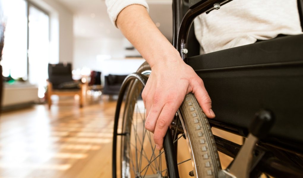 Nierozpoznawalna młoda niepełnosprawna kobieta na wózku inwalidzkim w domu w swoim salonie. Zbliżenie na jej rękę leżącą na kole.