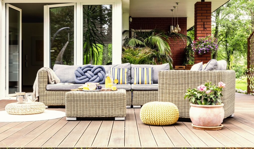 Stół ogrodowy z owocami i świeżym sokiem pomarańczowym stojący na drewnianym tarasie z roślinami, fotel i sofa z poduszkami