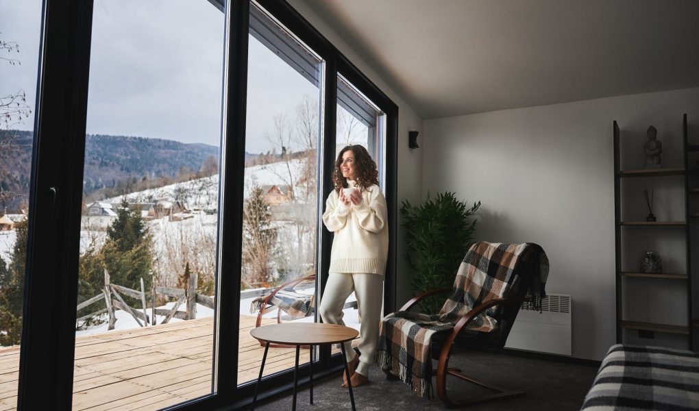 Kręcona młoda kobieta spędzająca weekend w nowoczesnym domu w górach. Szczęśliwa turystka patrząca przez panoramiczne okna w nowym domku.