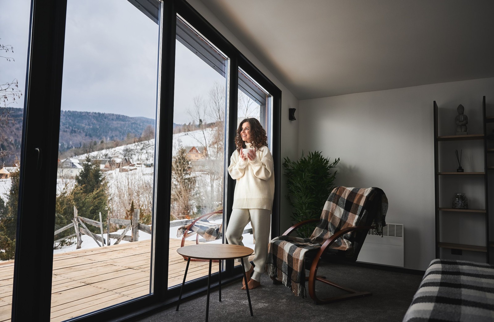 Kręcona młoda kobieta spędzająca weekend w nowoczesnym domu w górach. Szczęśliwa turystka patrząca przez panoramiczne okna w nowym domku.