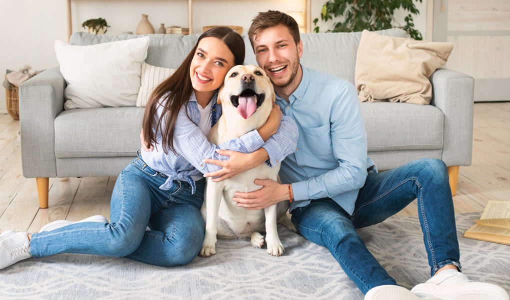 Koncepcja więzi, wspólnoty i przyjaźni. Portret uśmiechniętego małżeństwa przytulającego szczęśliwego psa siedzącego na podłodze w salonie w ich domu. Rodzina bawiąca się razem, patrząc w kamerę